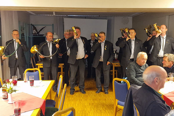 Männer spielen Jagdhörner im Anzug, stehend in einer Reihe. Im Hintergrund sind Tische mit Getränken und Blumen in einem Versammlungsraum, während einige Personen im Vordergrund sitzen.