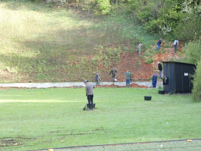 Personen sammeln auf einem grasbewachsenen Schießplatz Projektilreste ein. Einige tragen Eimer und Rechen, während sie eine Böschung erklimmen. Ein schwarzes Gebäude steht rechts.