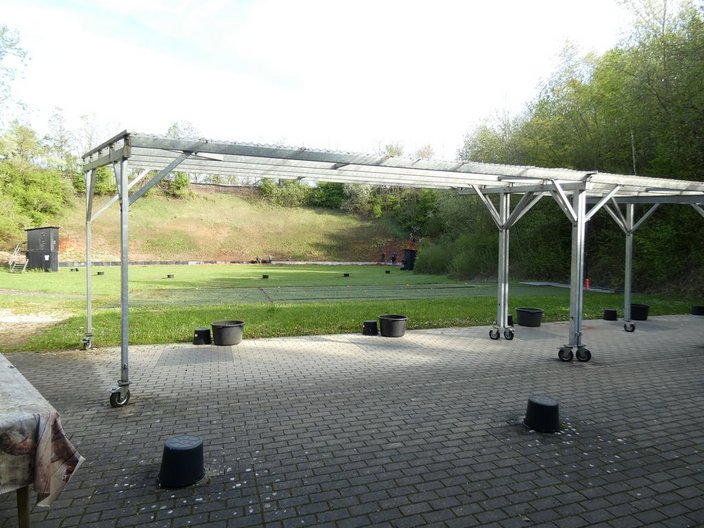 Metallkonstruktion mit Rädern steht auf gepflastertem Platz; viele schwarze Eimer liegen verstreut; Grasfläche und Bäume im Hintergrund, sonnige Umgebung.