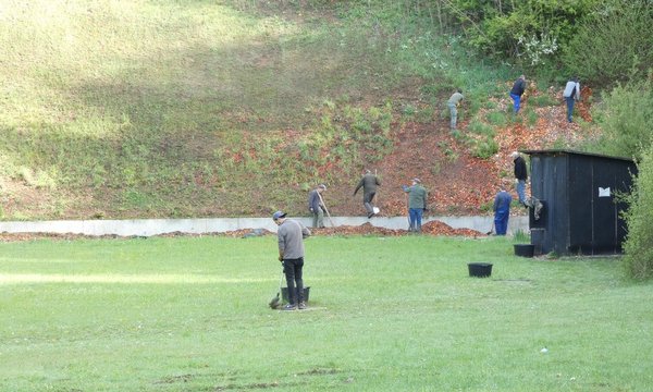 Personen sammeln auf einem grasbewachsenen Schießplatz Projektilreste ein. Einige tragen Eimer und Rechen, während sie eine Böschung erklimmen. Ein schwarzes Gebäude steht rechts.